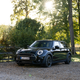 A black and gold 2023 Mini Cooper Clubman is parked on gravel in front of some trees.